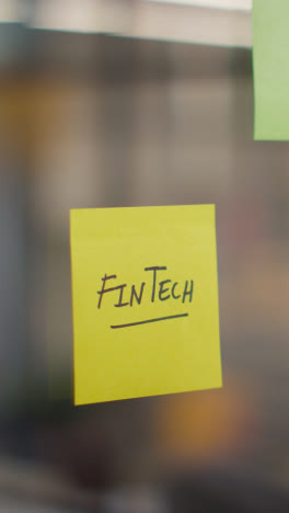 Vertical-Video-Close-Up-Of-Woman-Putting-Sticky-Note-With-Fintech-Written-On-It-Onto-Transparent-Screen-In-Office